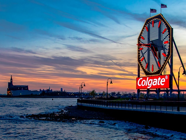 pajūris, „Colgate“ logotipas fone po dideliu laikrodžiu su raudonomis rodyklėmis