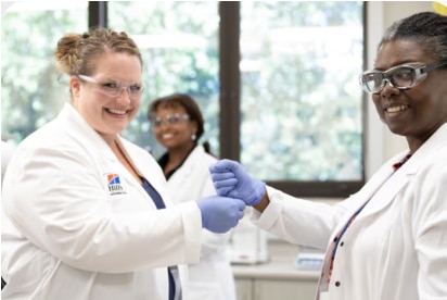 Du mokslininkai, vilkintys laboratorinius chalatus ir pirštines, šypsosi ir spaudžia vienas kitam rankas laboratorijos aplinkoje.